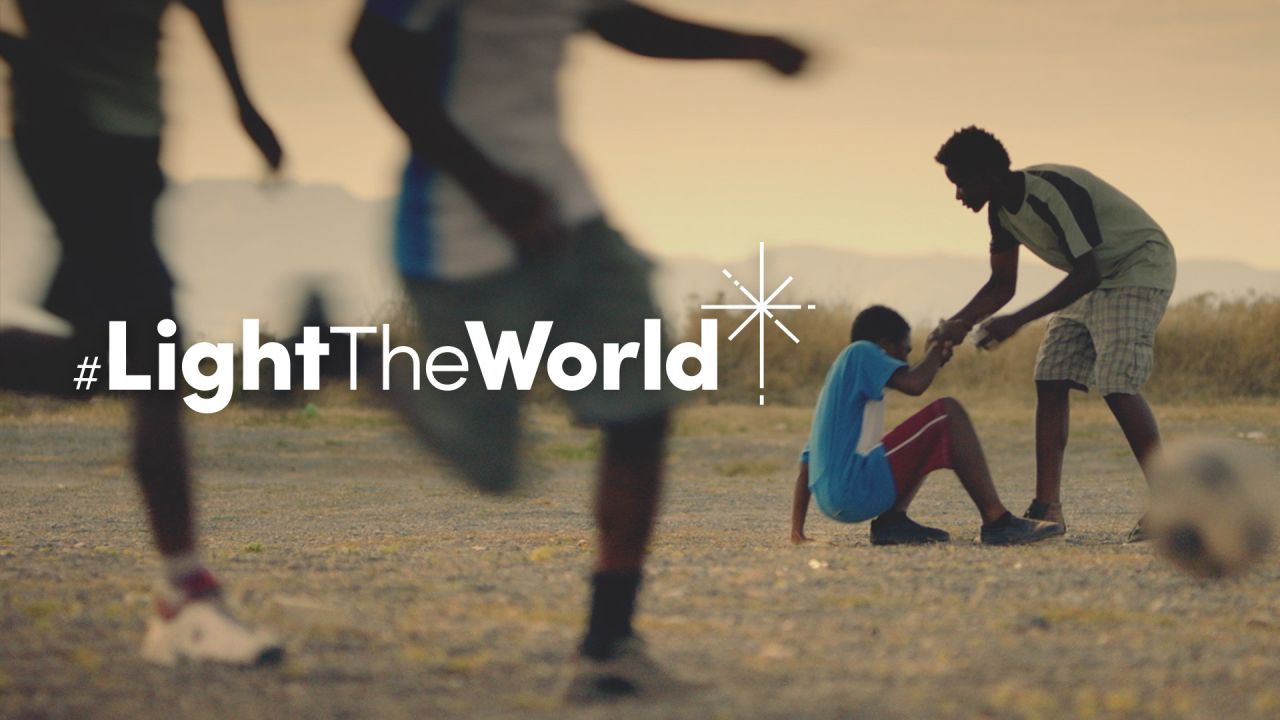 Um menino ajuda outro a levantar-se depois deste ter caído a jogar futebol ― do vídeo Seja a Luz do Mundo
